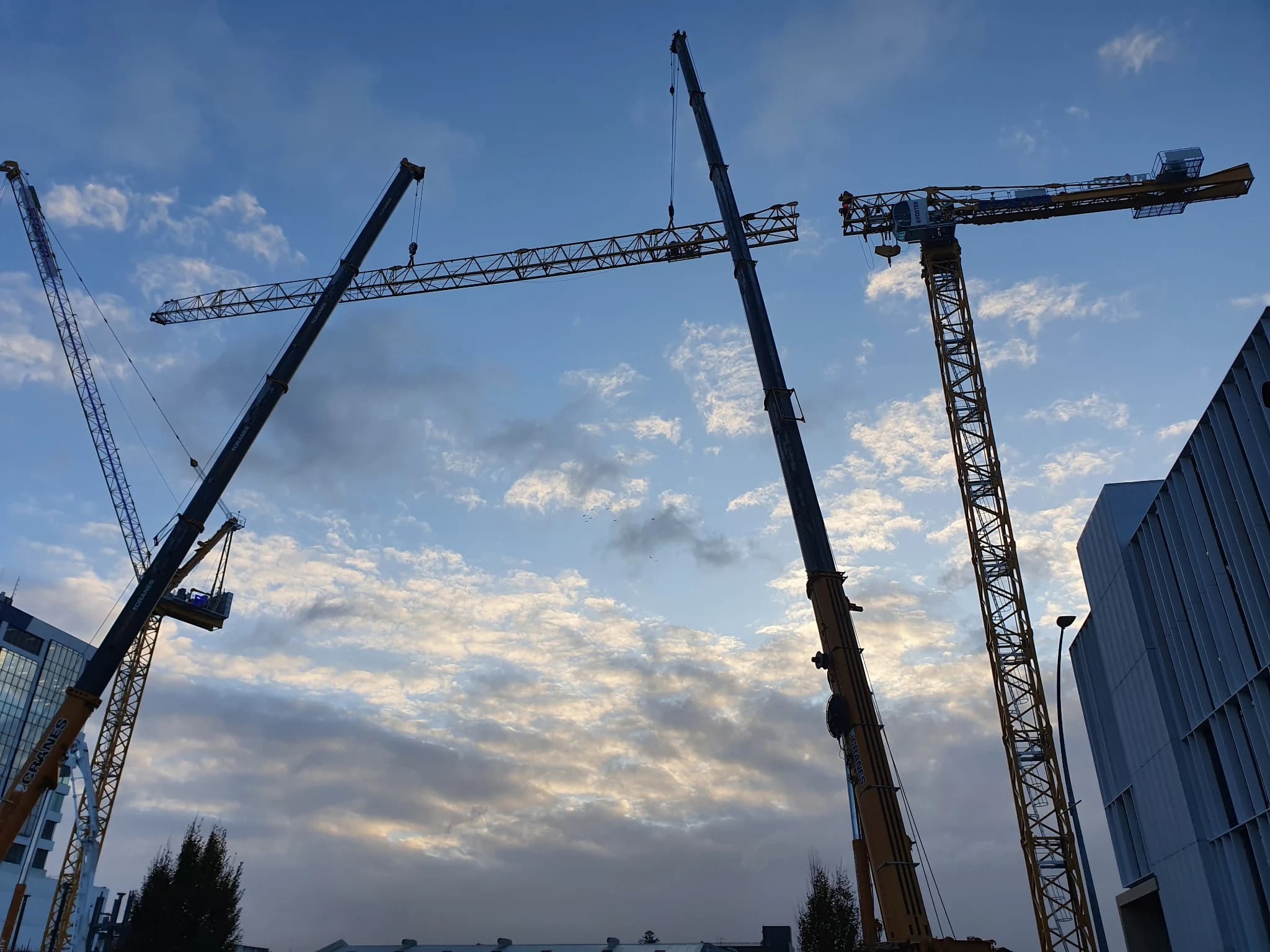 Two tower cranes and a mobile crane rising against a bright, cloud-streaked sky, seen from street level.