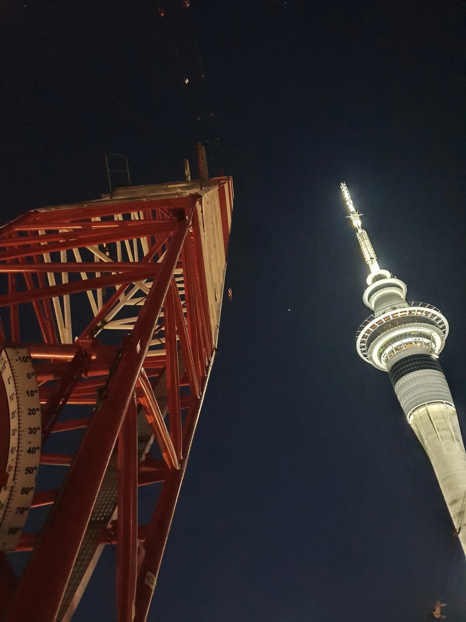 A red-lit crane mast at night beside the lit Sky Tower.