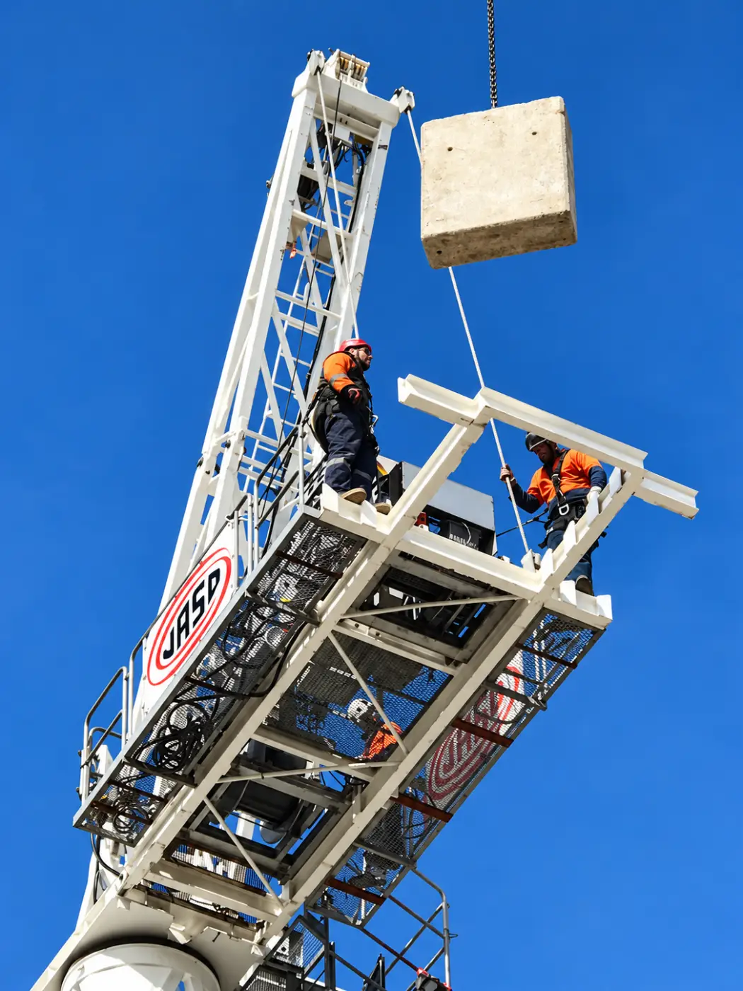 Crew on a crane platform high above, a concrete counterweight hanging against a blue sky.