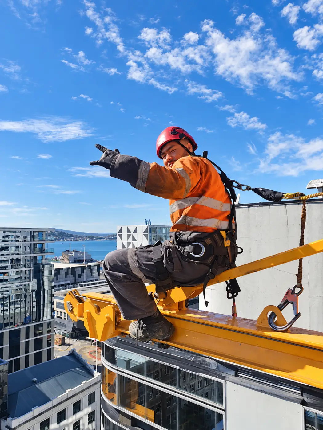 A rigger in a harness on a yellow crane above Auckland, pointing across the city.